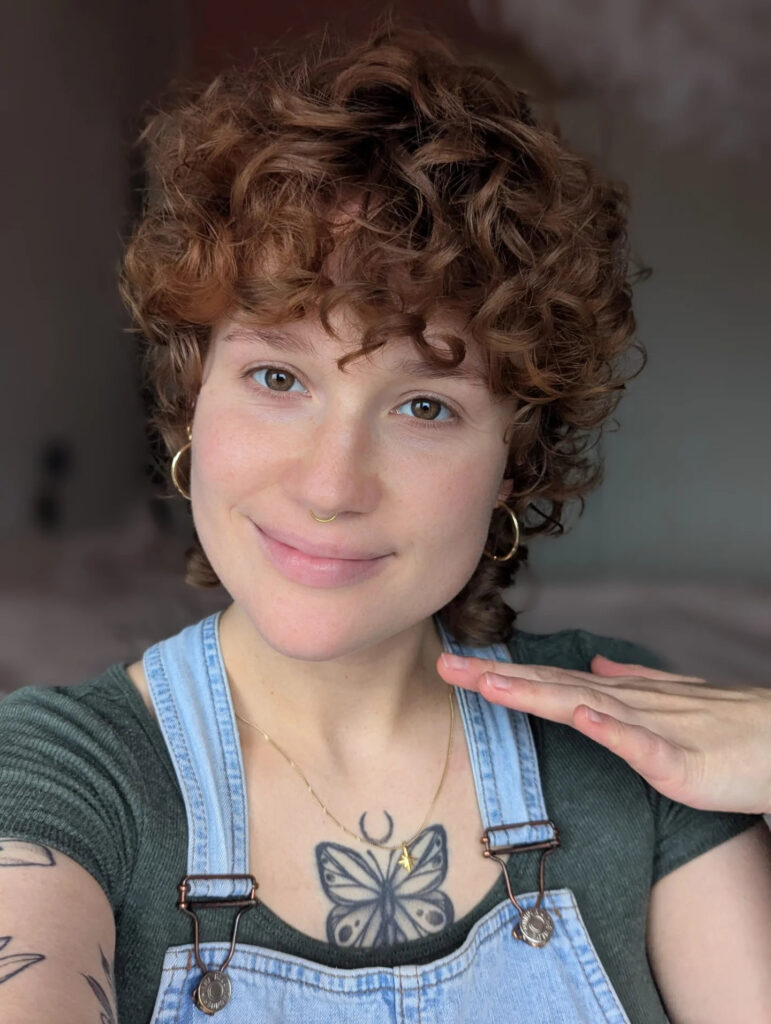A woman with short curly auburn hair and a soft curly fringe is smiling at the camera.