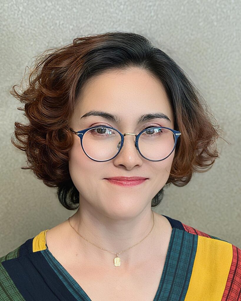A woman wearing blue glasses with a chin-length bob featuring dark roots, auburn ends, and soft, polished waves.