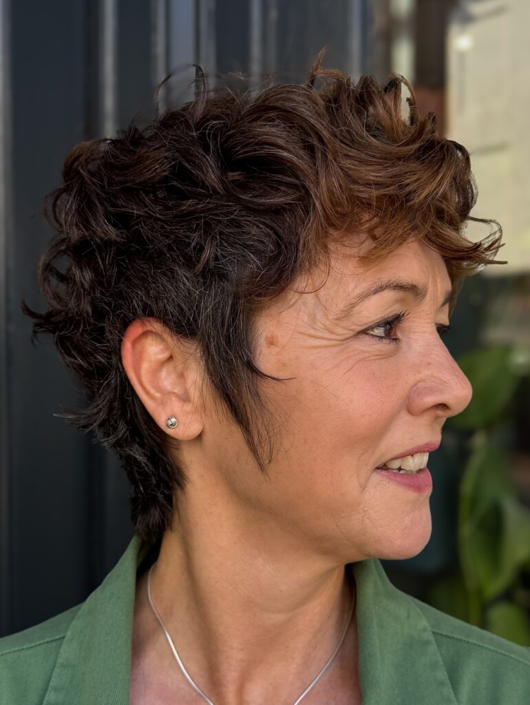 A side profile of a woman with a dark brunette textured pixie cut featuring soft curls and subtle brown highlights.