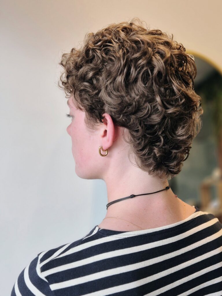 A side and back view of a woman with a tapered ash-brown curly hairstyle and gold hoop earrings.
