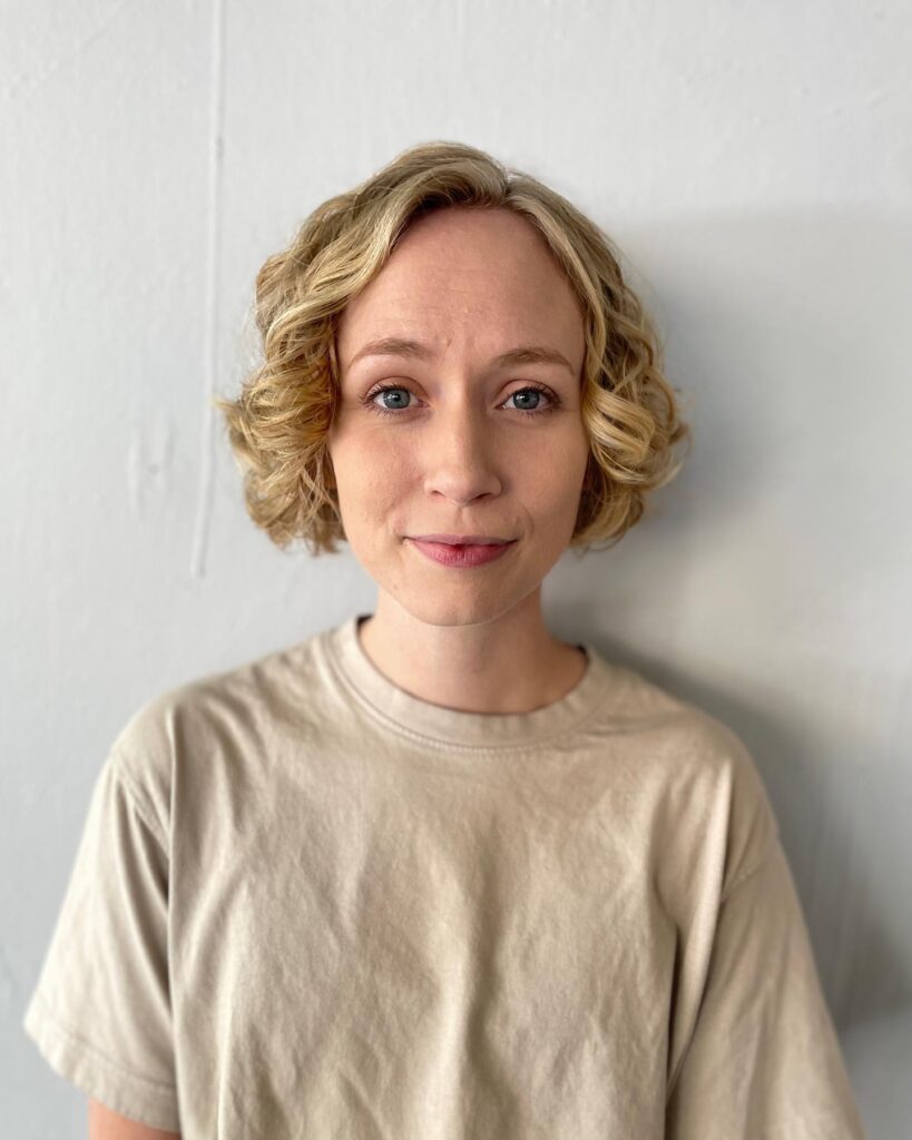 A woman with a chin-length honey blonde bob styled in soft, polished waves with a side part.