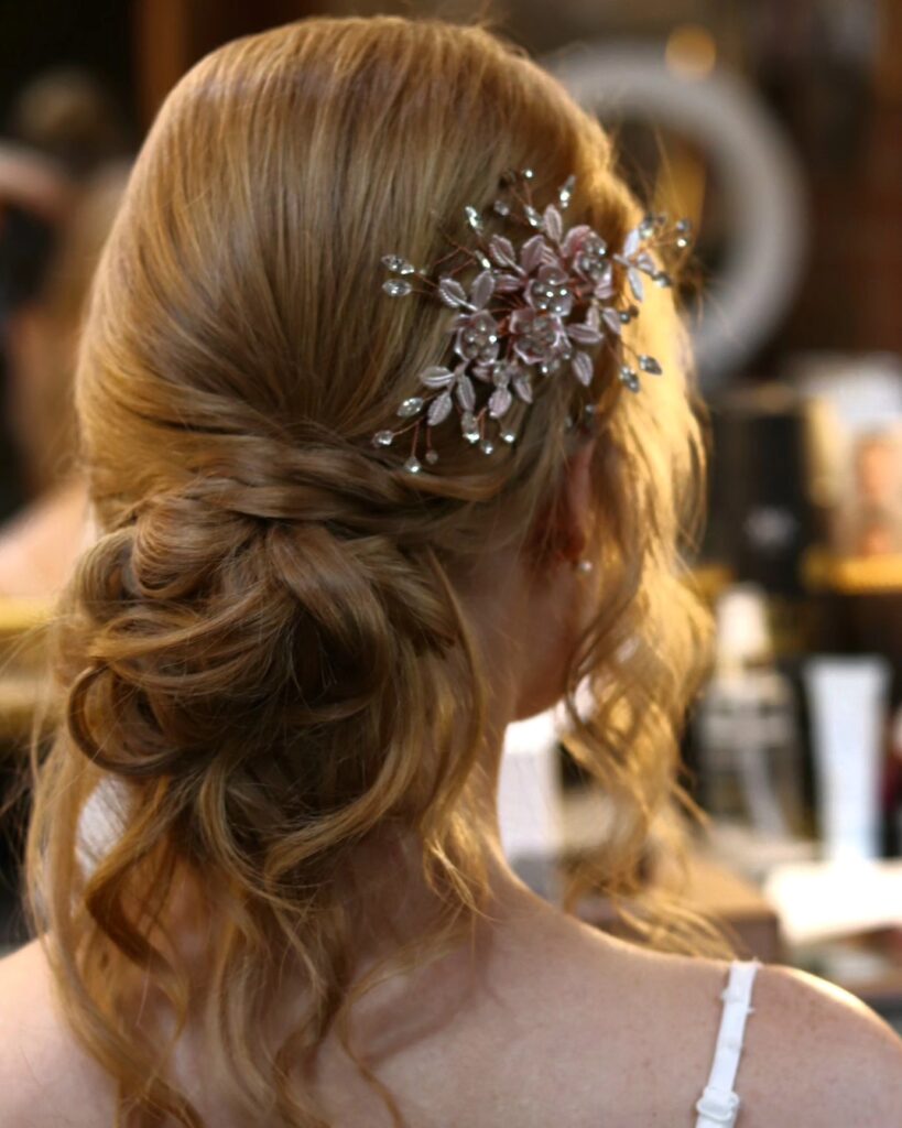 A close-up from behind of a woman with a messy low bun hairstyle decorated with a crystal flower hair clip and loose wavy curls.