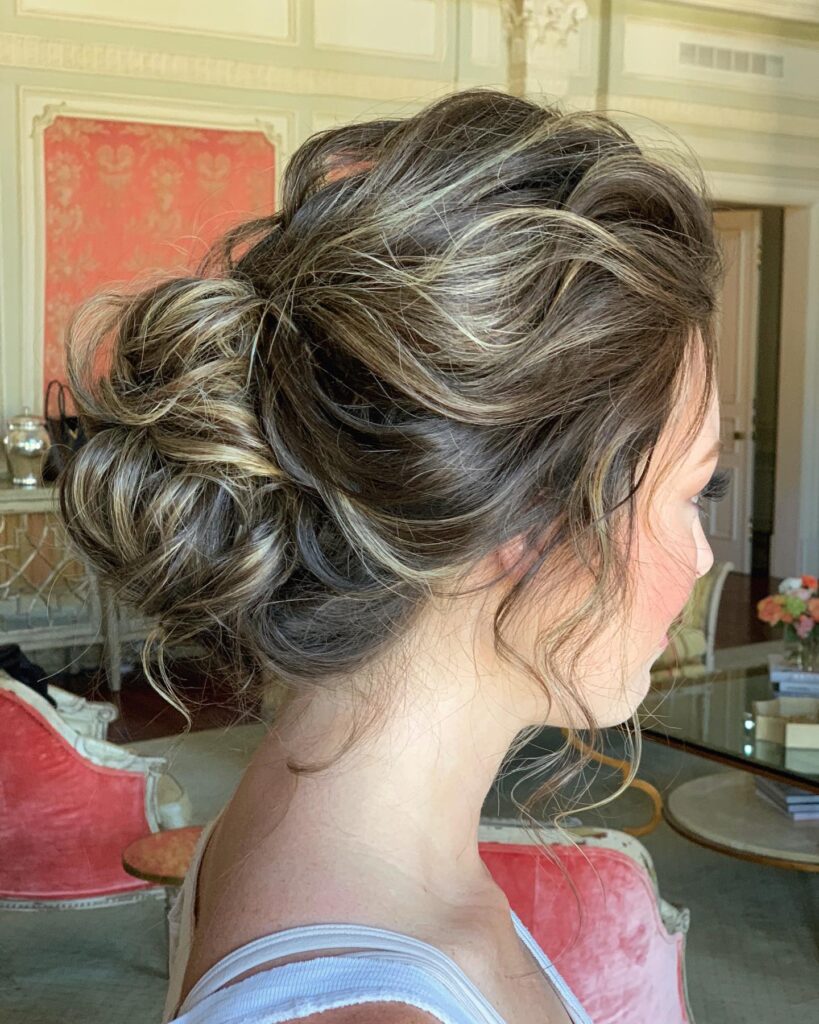 A side profile of a woman with a voluminous messy bun featuring dark brown hair and bright blonde highlights with wavy face-framing pieces.