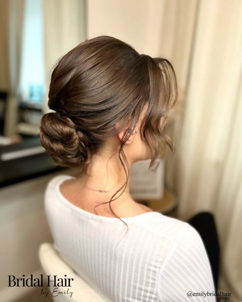 A side profile of a woman with a smooth brunette low coiled bun and soft wavy face-framing tendrils.