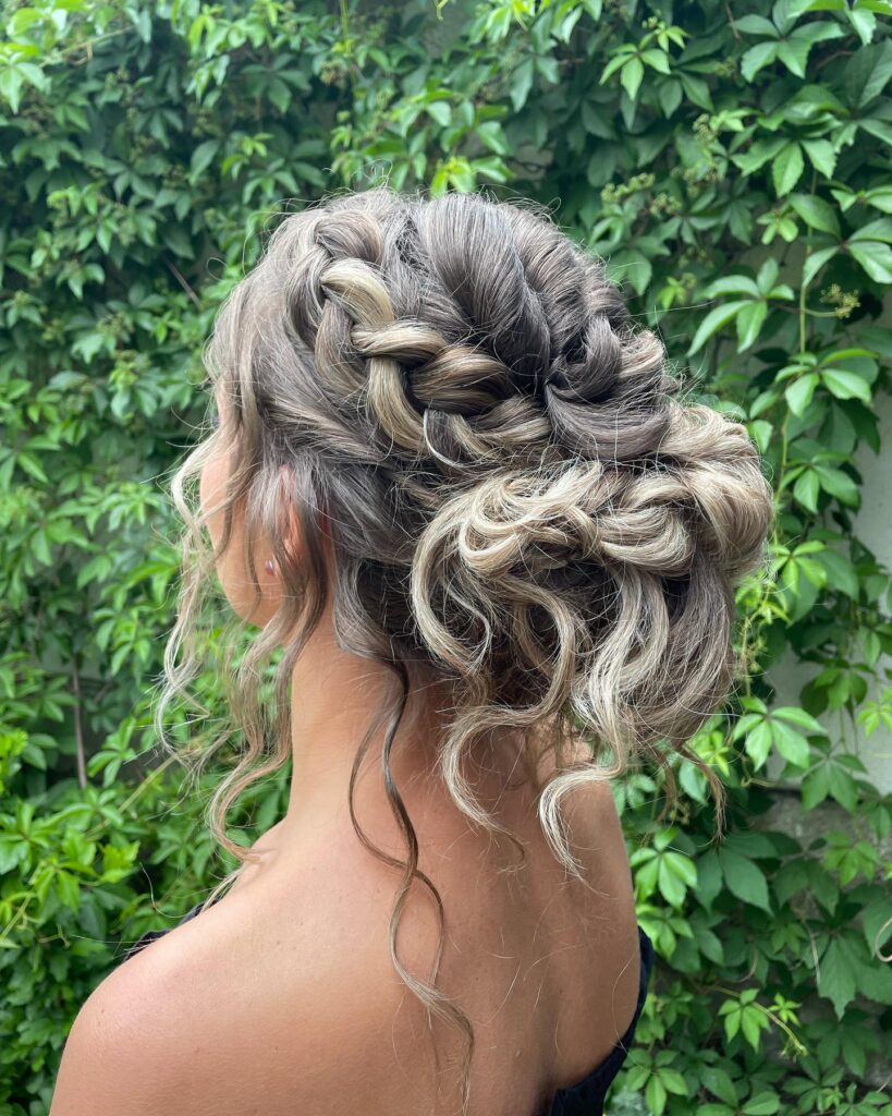 A side view of a woman featuring a thick Dutch braid that transitions into a messy low bun with loose ash blonde curls.