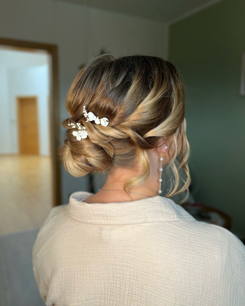 A woman seen from behind with a twisted honey-blonde chignon decorated with small white floral hairpins.