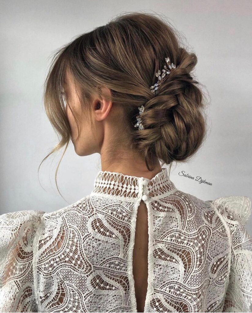 A side profile of a woman with an ash brown braided low bun decorated with small crystal hairpins and soft face-framing pieces.