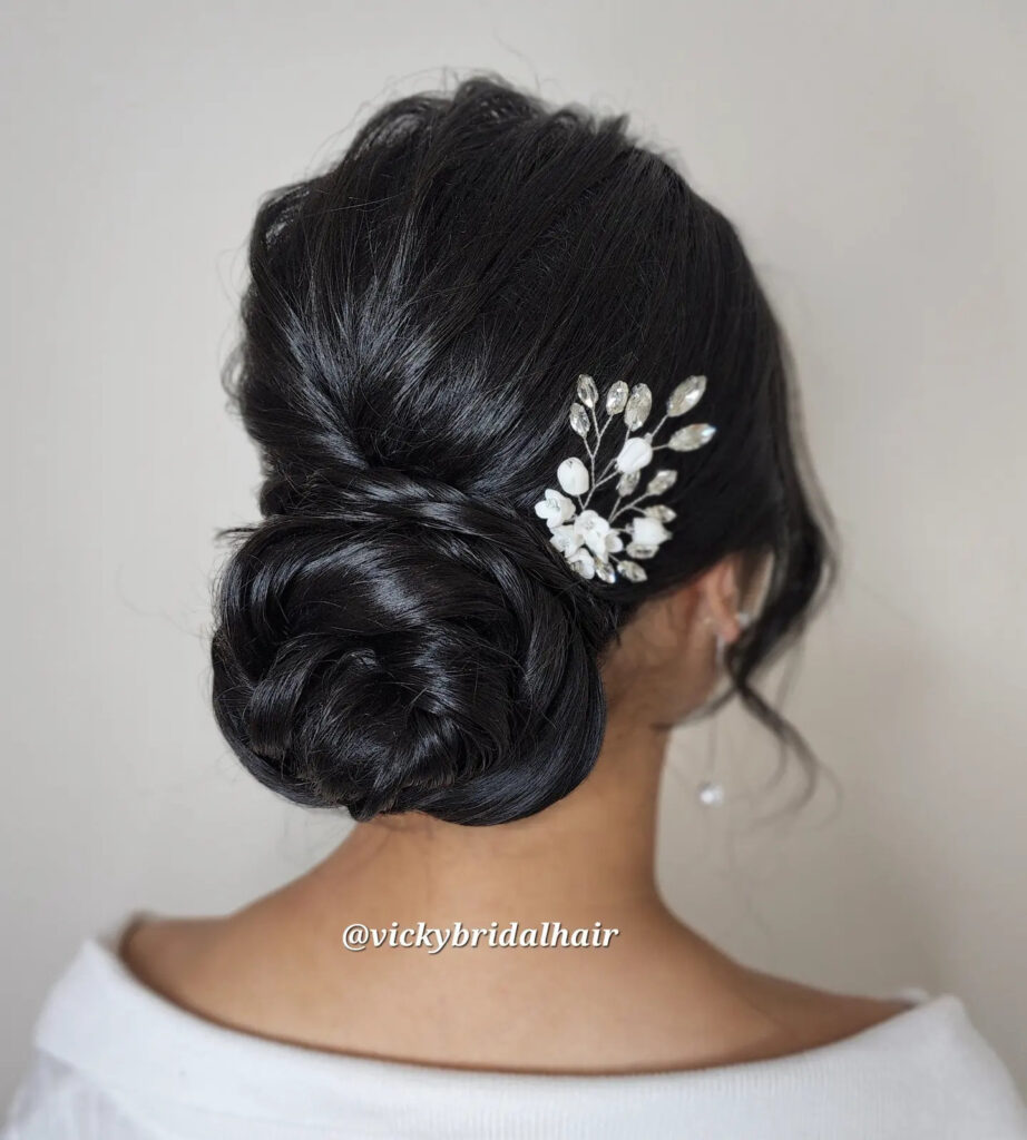 A woman from behind with glossy black hair styled into an intricate rosette low bun, decorated with a white floral crystal hairpiece.