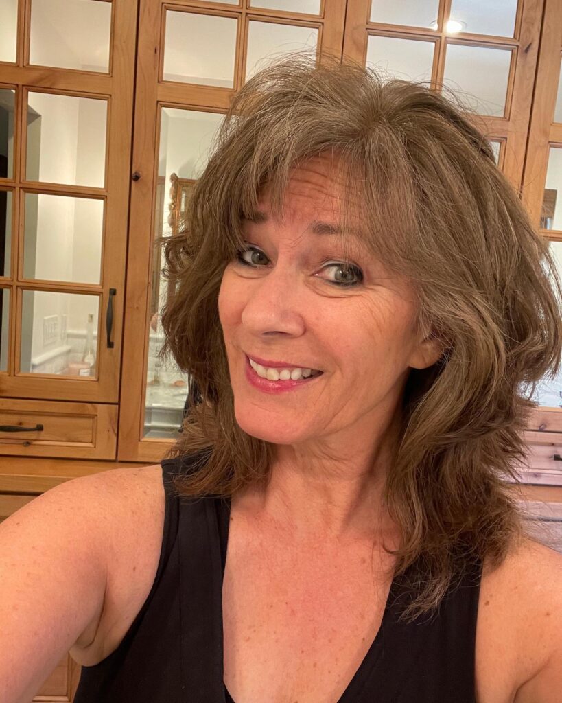 A smiling woman with a voluminous shoulder-length shaggy haircut in ash brown with wispy bangs.