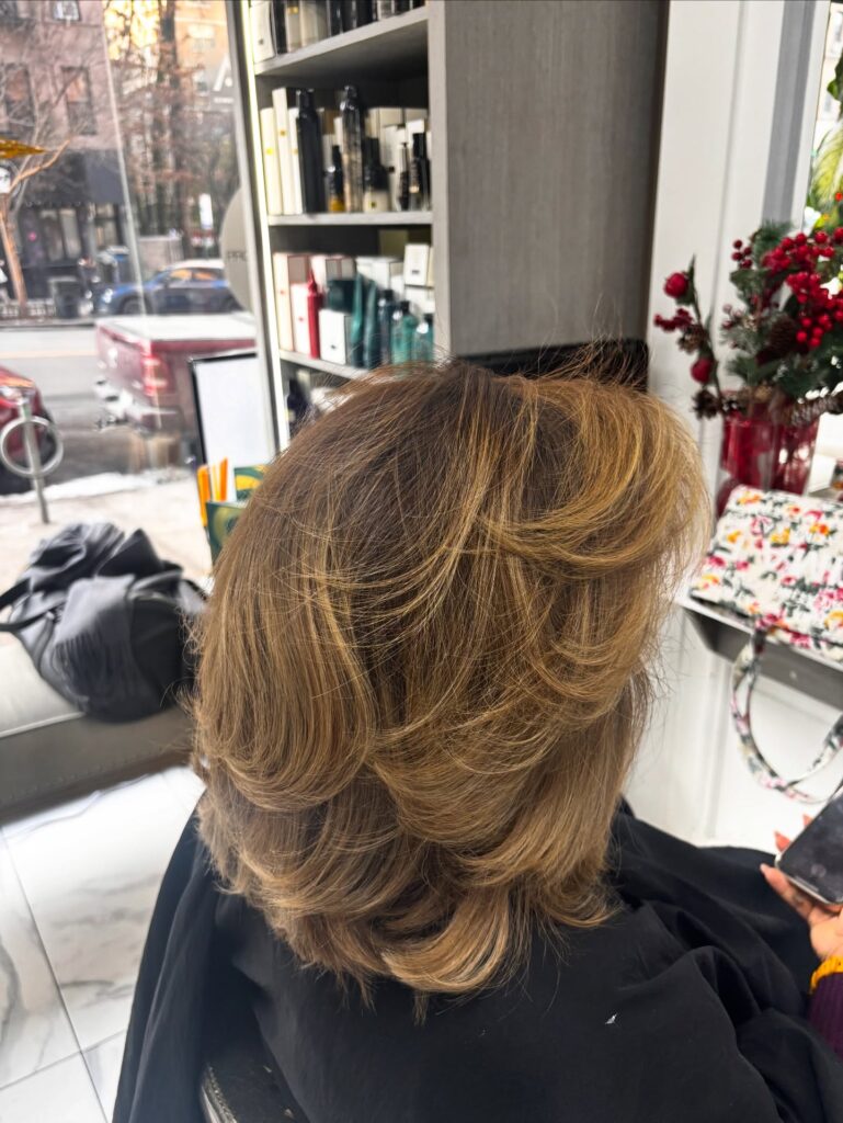A side view of a woman with shoulder-length layered honey-blonde hair featuring voluminous feathered layers and a blowout style.