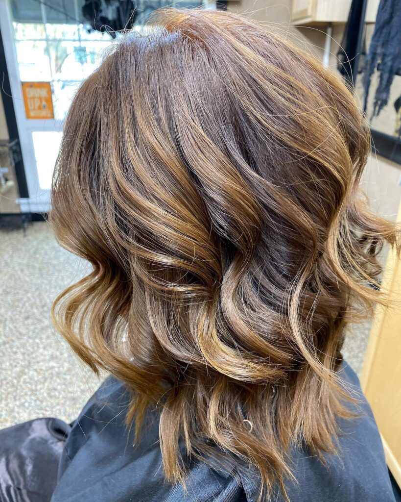A back view of a woman with shoulder-length layered hair featuring honey blonde balayage and beachy waves.