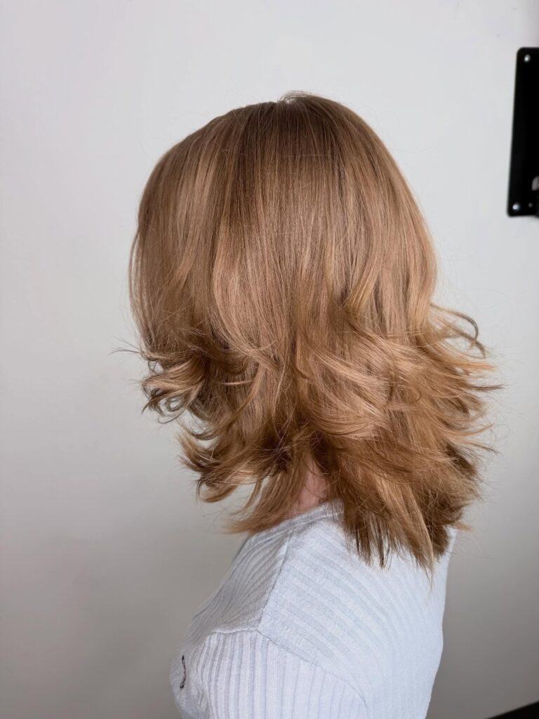 A side and back view of a woman with shoulder-length layered sandstone-blonde hair featuring soft feathered ends.