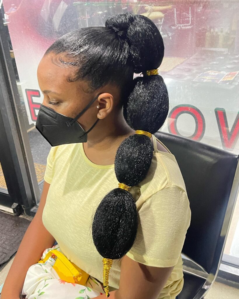 A woman wears a high black bubble ponytail accented with decorative gold wire wraps between each voluminous section.