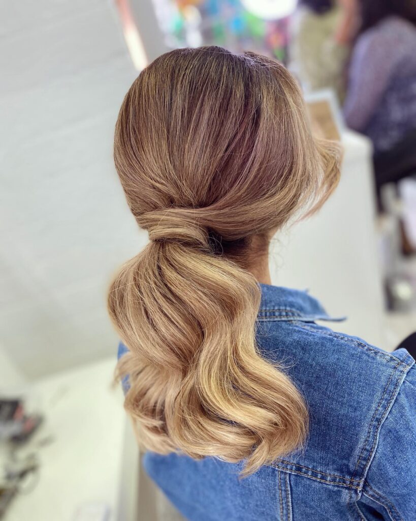 A woman with wavy honey-blonde hair gathered into a soft low ponytail with a thick hair wrap and face-framing strands.