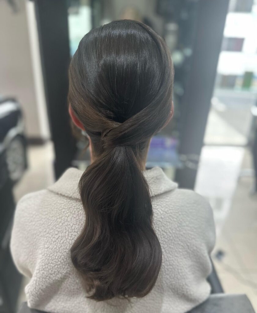 Rear view of a woman with a low brunette ponytail featuring an intricate twisted wrap at the base and soft wide waves.