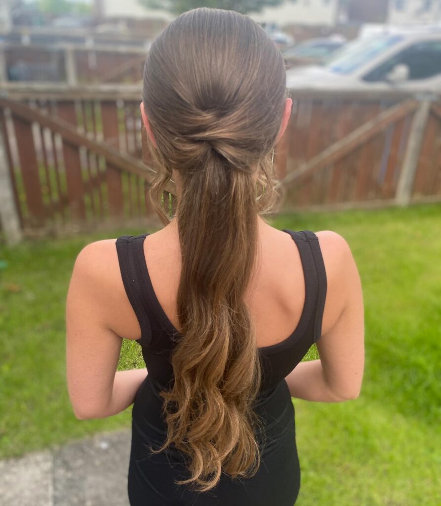 Rear view of a woman with long, wavy ash-brown hair styled in a low ponytail with a decorative tucked hair wrap at the base.