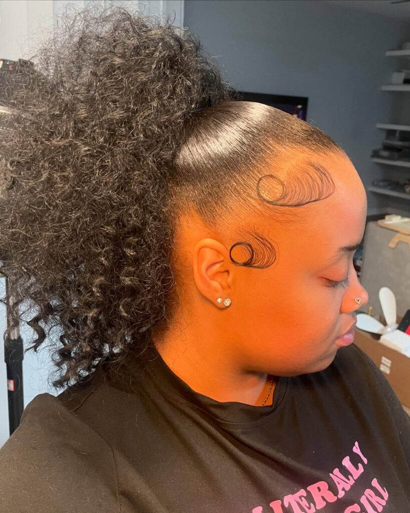 A woman with a sleek high ponytail featuring voluminous natural curls and intricate circular swirls of baby hairs along her forehead and temple.