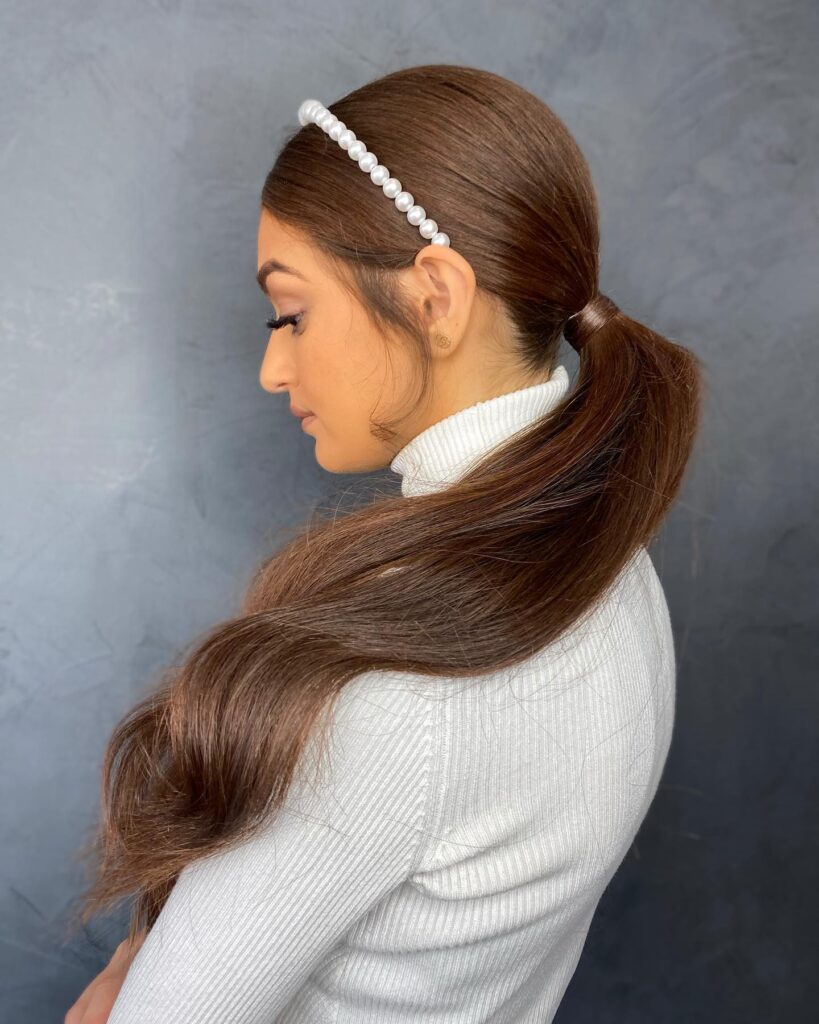 A woman in profile wearing a sleek, low chestnut brown ponytail accessorized with a white pearl headband and soft, wispy side curls.