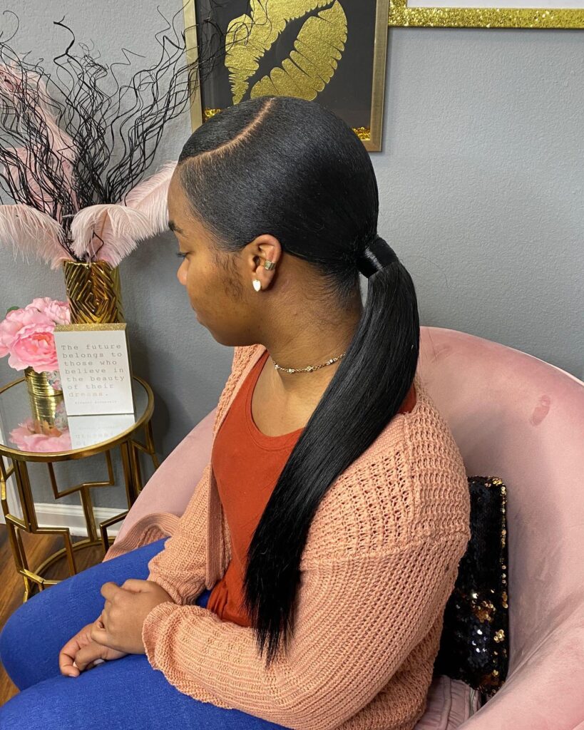 A woman in profile wearing a sleek side-parted low ponytail with a straight black texture and a polished hair wrap.