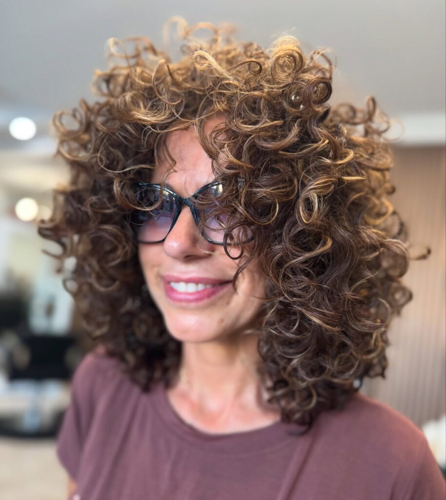 A woman with voluminous shoulder-length brown curls featuring warm caramel highlights.