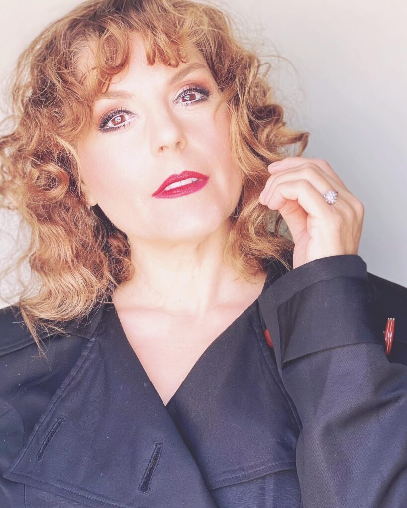 A woman with medium-length honey-copper hair styled in a bouncy curly shag with soft bangs.