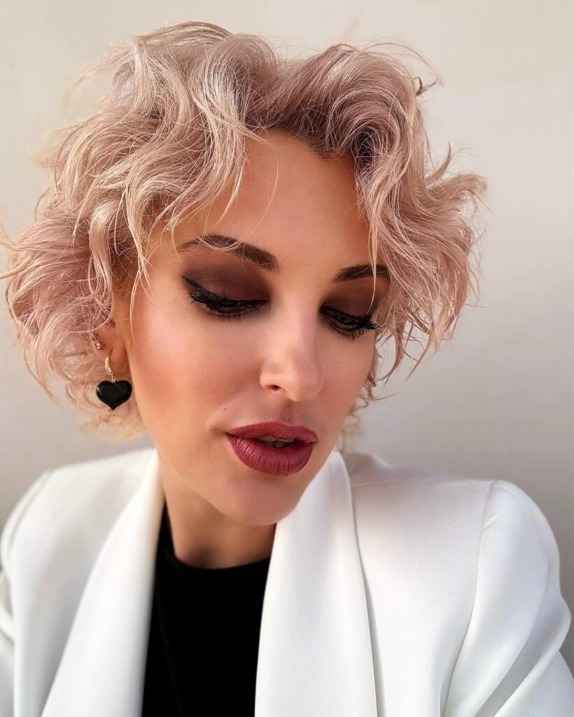 A woman with a chin-length rose gold bob styled in soft, tousled curls and wispy layers.