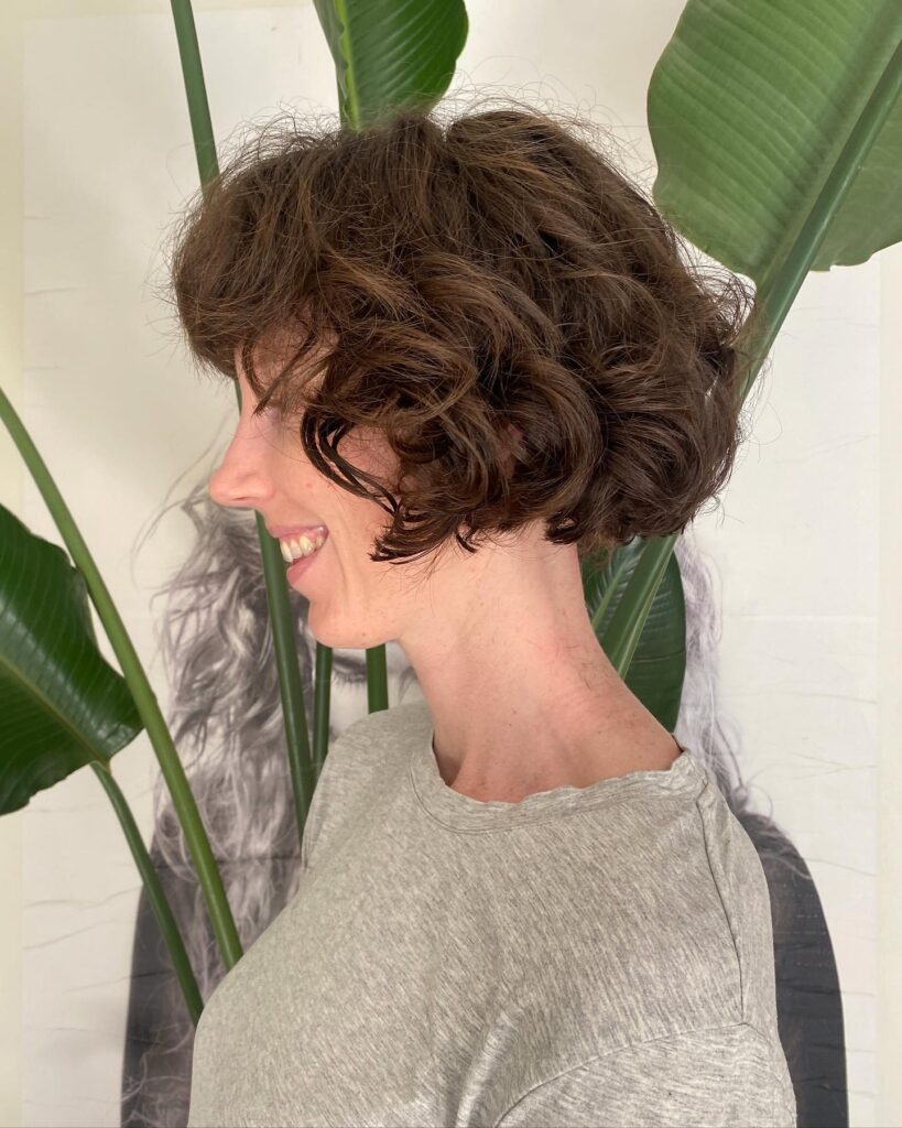 A woman seen from the side with a chocolate brown, chin-length bob styled in soft, textured curls.