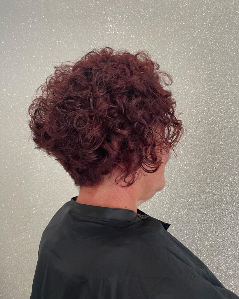 A woman seen from the side with a graduated bob featuring tight mahogany red curls and a stacked back.