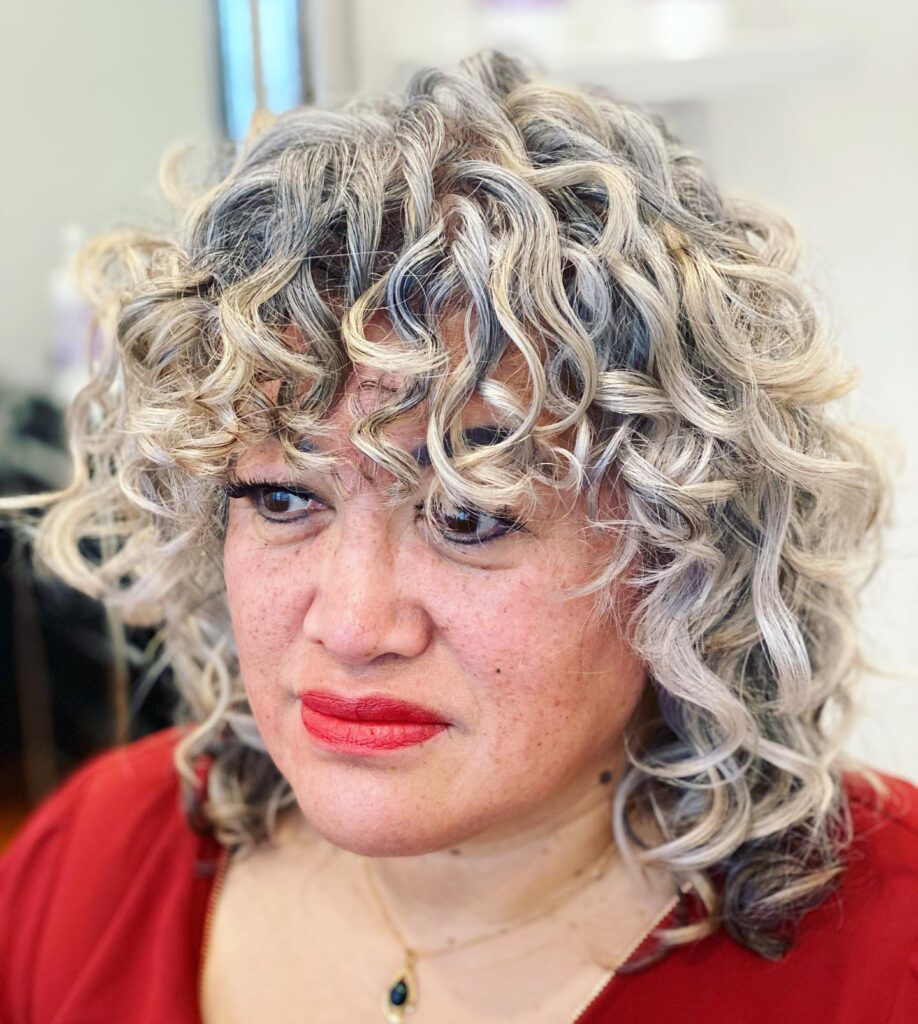 A woman with shoulder-length silver and charcoal curls featuring a bouncy curly fringe.