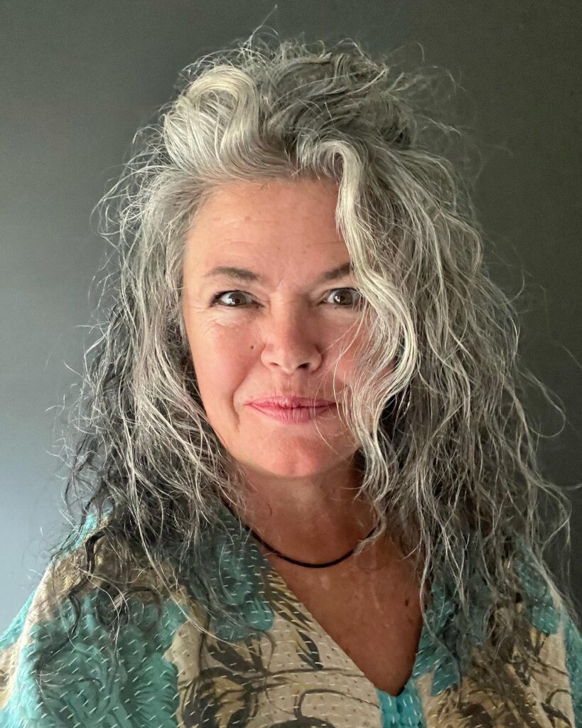 A woman with long salt-and-pepper hair styled in a voluminous curly shag with silver face-framing layers.
