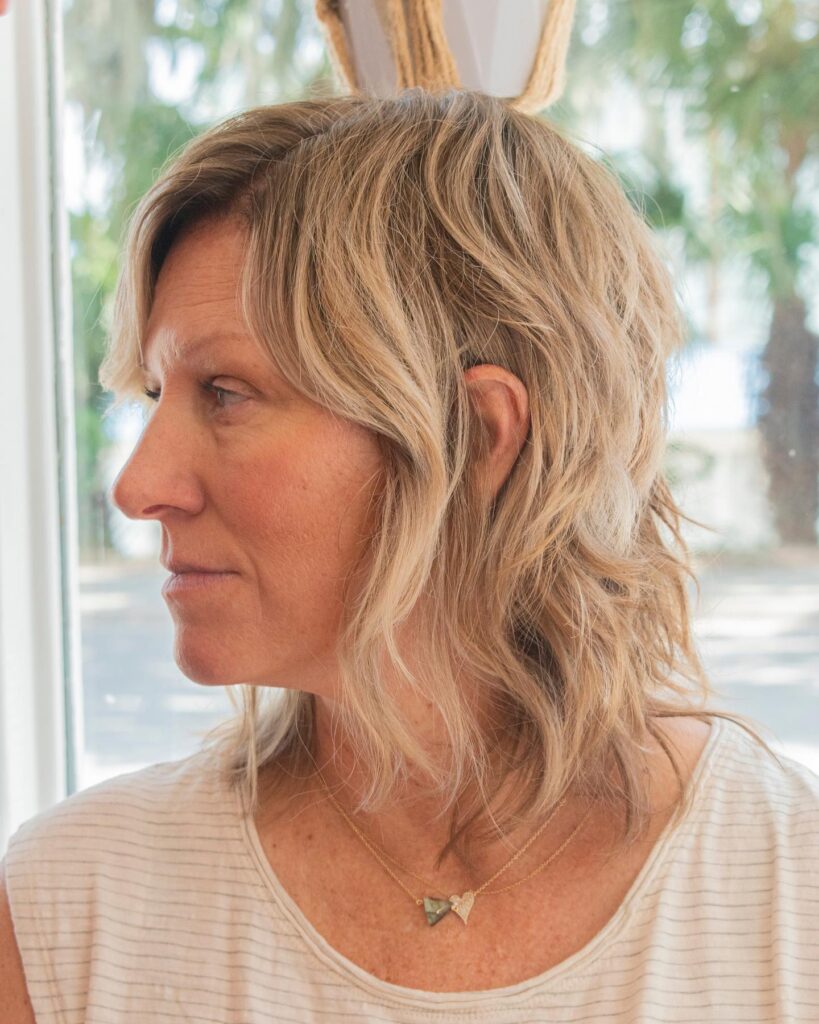 A side profile of a woman with a sandy blonde medium-length shaggy haircut and wavy layers.