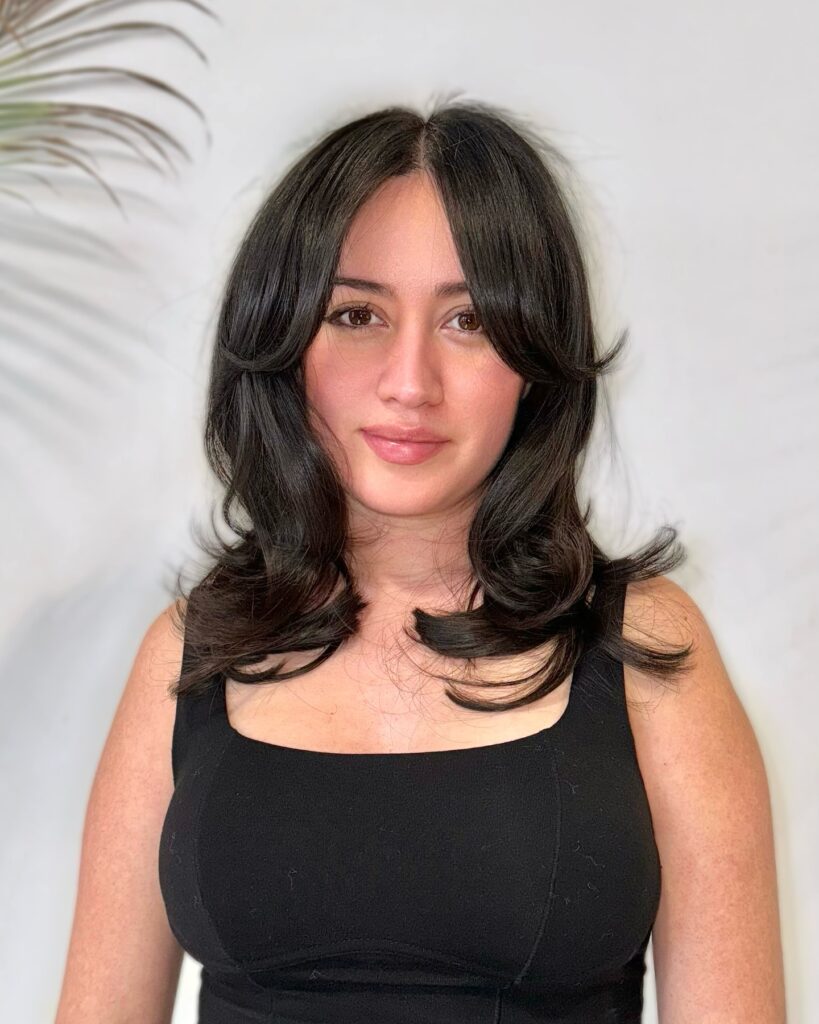 A woman with glossy dark brown medium length hair styled in voluminous butterfly layers and face framing curtain bangs.