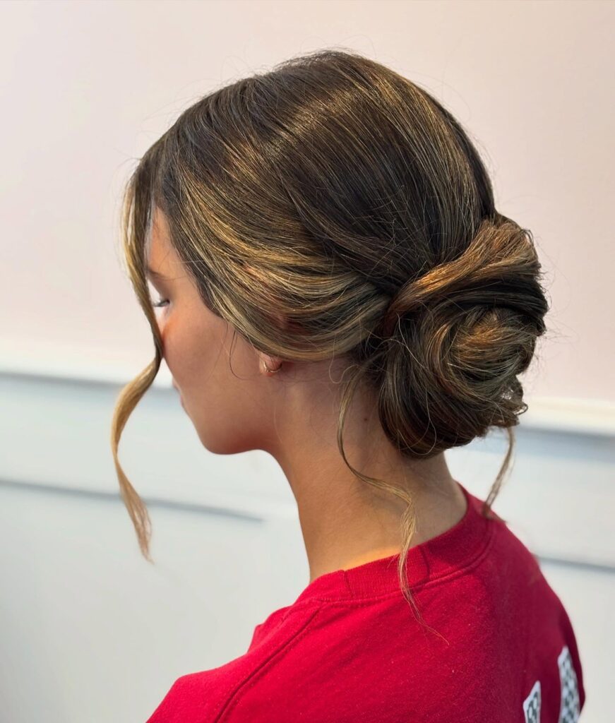 A woman with highlighted brown hair styled in a low twisted bun with loose wavy strands framing her face.