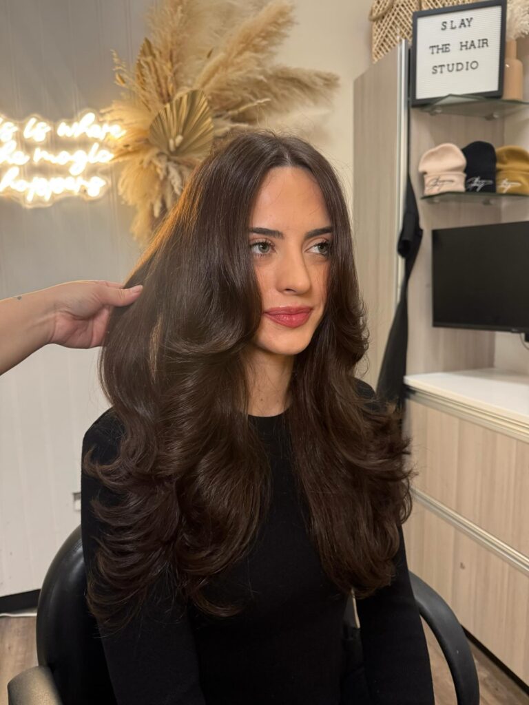 A woman with long voluminous dark brown hair styled in dramatic butterfly layers with bouncy curled ends.