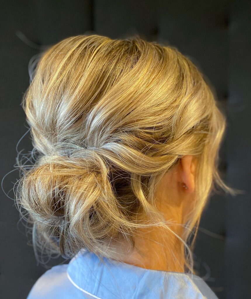 A back view of a woman with multi tonal blonde hair styled in a voluminous and textured messy low bun.