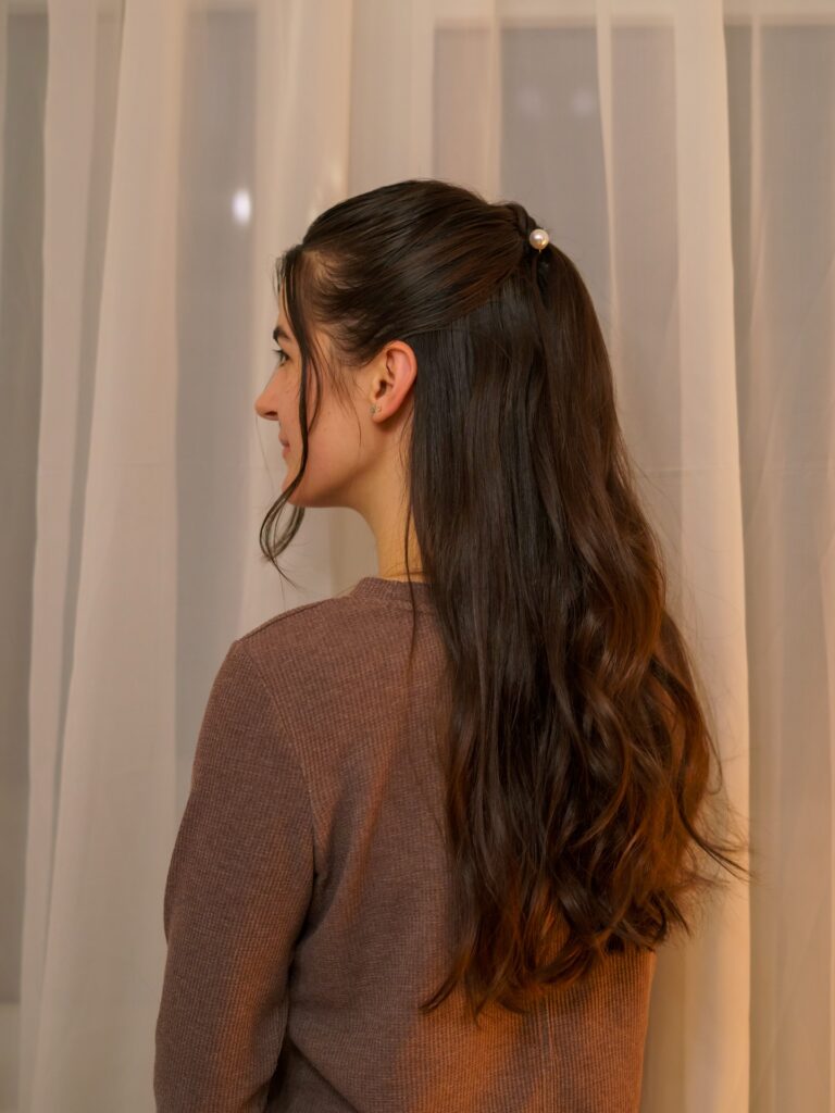 A side profile view of a woman with long wavy brunette hair styled in a half up look secured with a single pearl hair pin.
