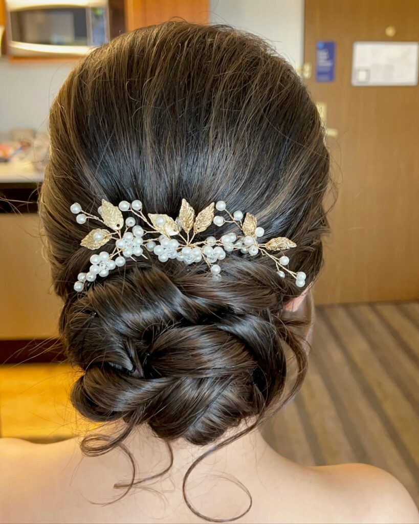 A woman with dark brown hair wears a low twisted chignon decorated with a gold leaf and pearl hair accessory.