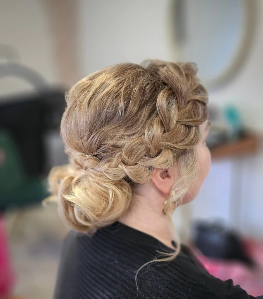 A woman with intricate, blonde-highlighted woven hair is styled in a loose, low bun with pieces framing her face.