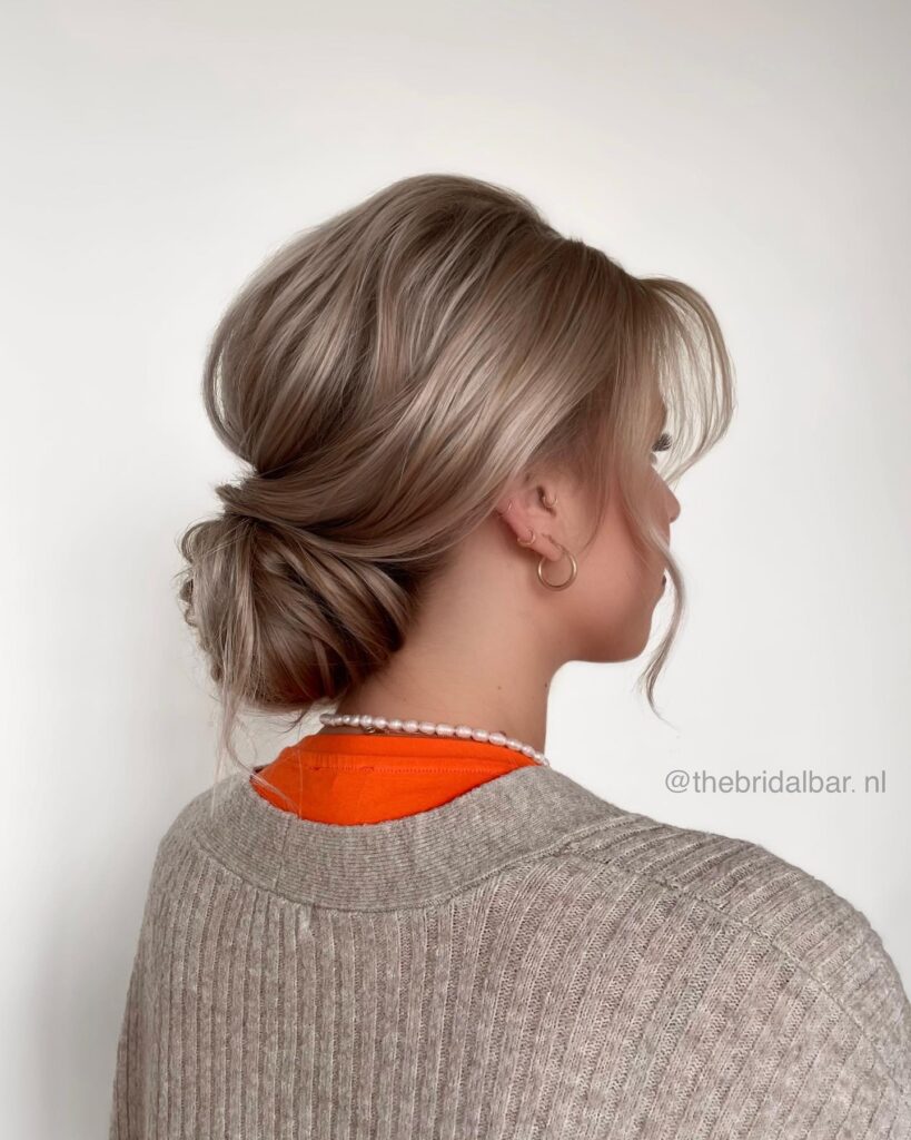 A woman with ash blonde hair wears a smooth low tucked bun with soft face-framing fringe.