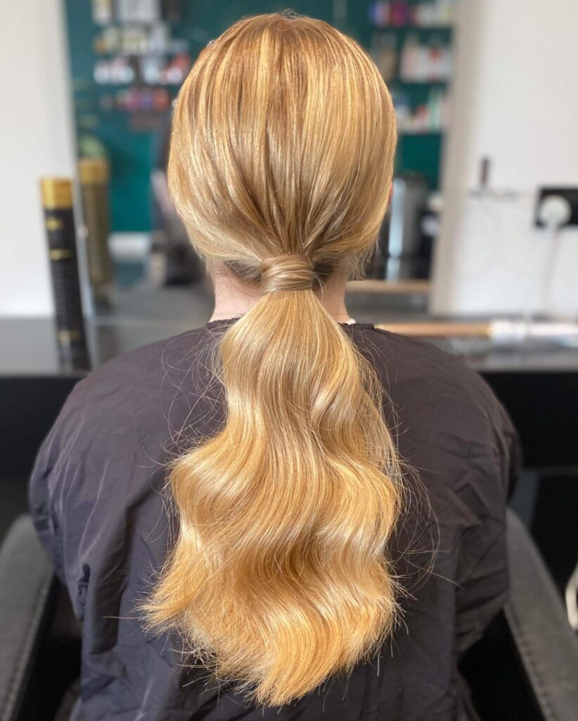 Long blonde hair styled in a low ponytail with soft loose waves flowing down the back.