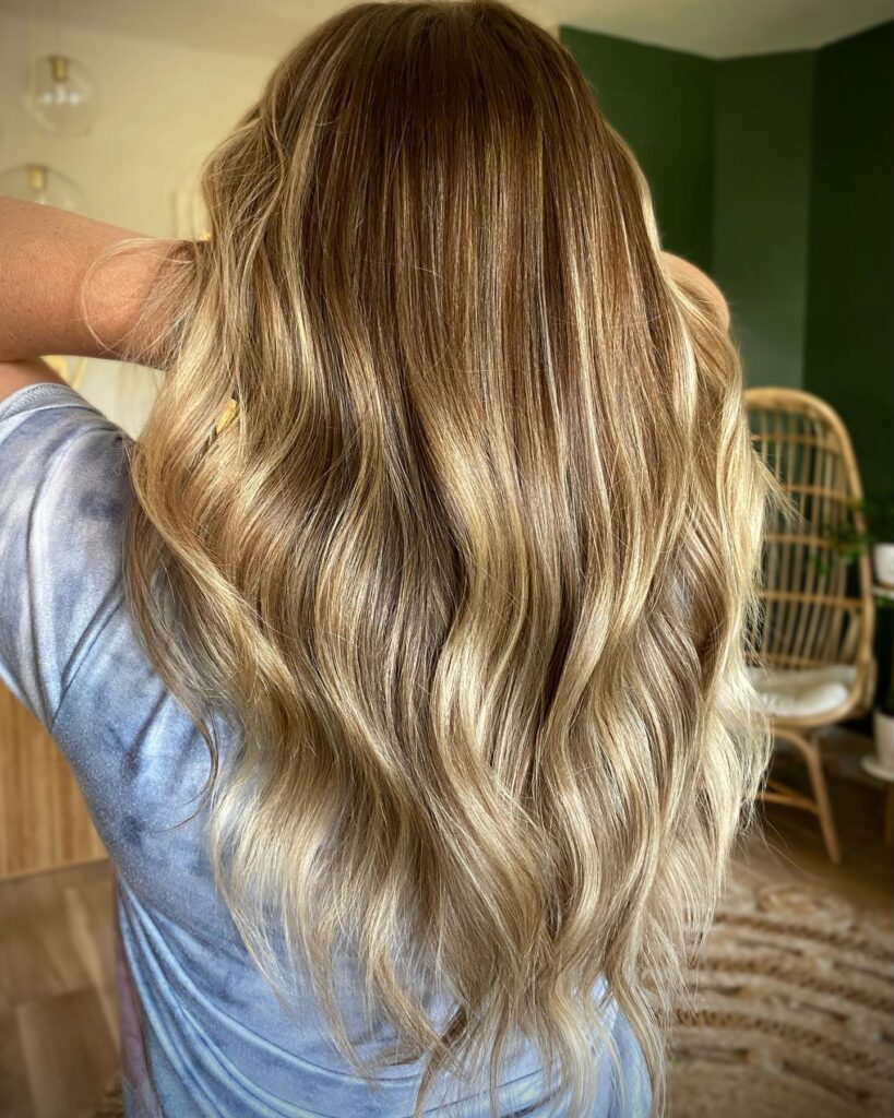 The back view of a woman with long wavy hair featuring a blend of warm honey and golden blonde highlights.