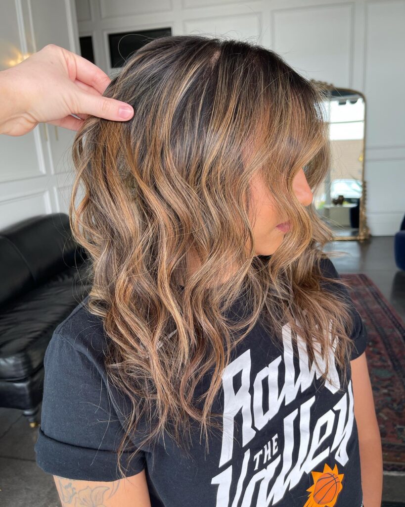 A woman with medium length wavy dark hair featuring warm toffee highlights and textured layers.