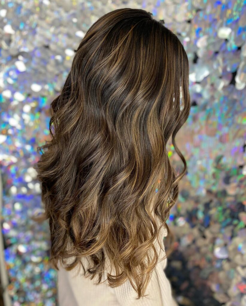 A woman with long wavy dark brown hair featuring bright caramel highlights and soft layers against a shimmering silver background.