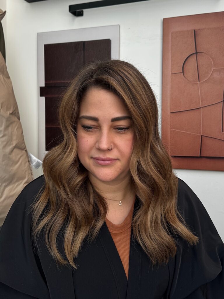 A woman with medium length wavy mocha brown hair and soft honey blonde highlights looking down slightly.