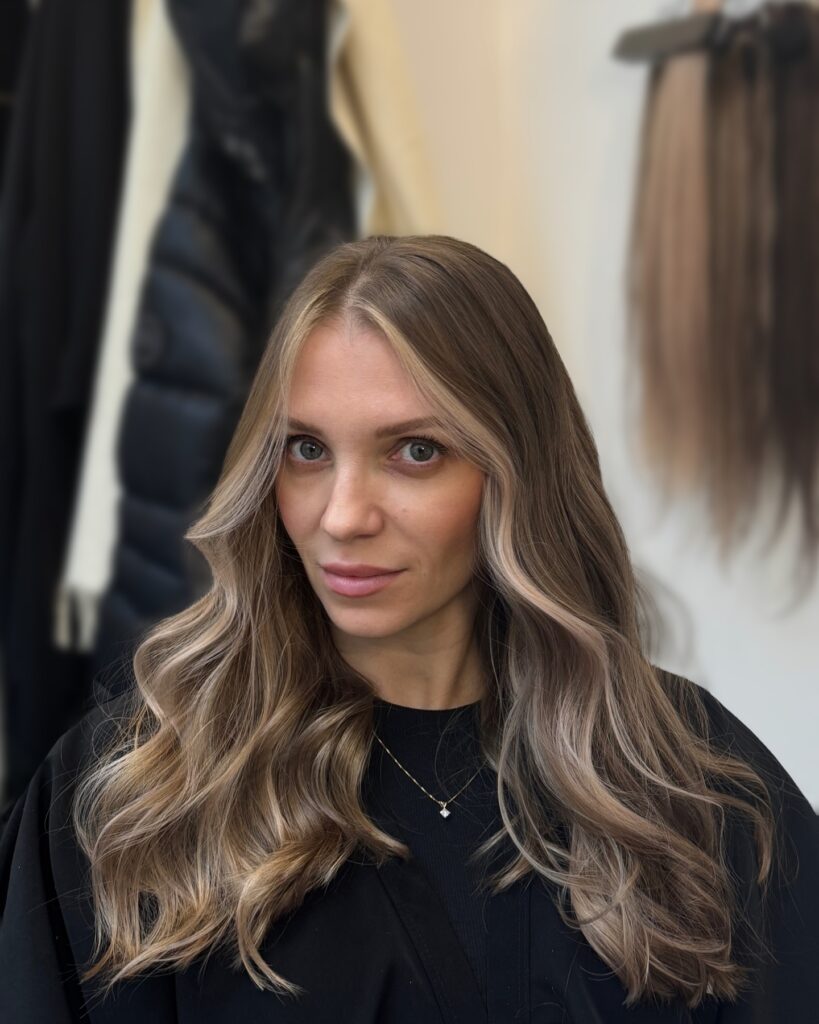 A woman with long wavy ash blonde hair and subtle dark roots looking forward with soft face framing highlights.