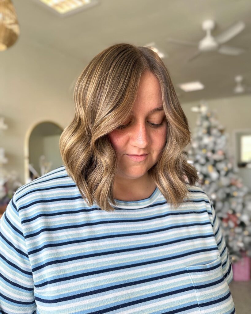 A woman with a shoulder length lob hairstyle featuring sandy blonde highlights and soft textured waves.