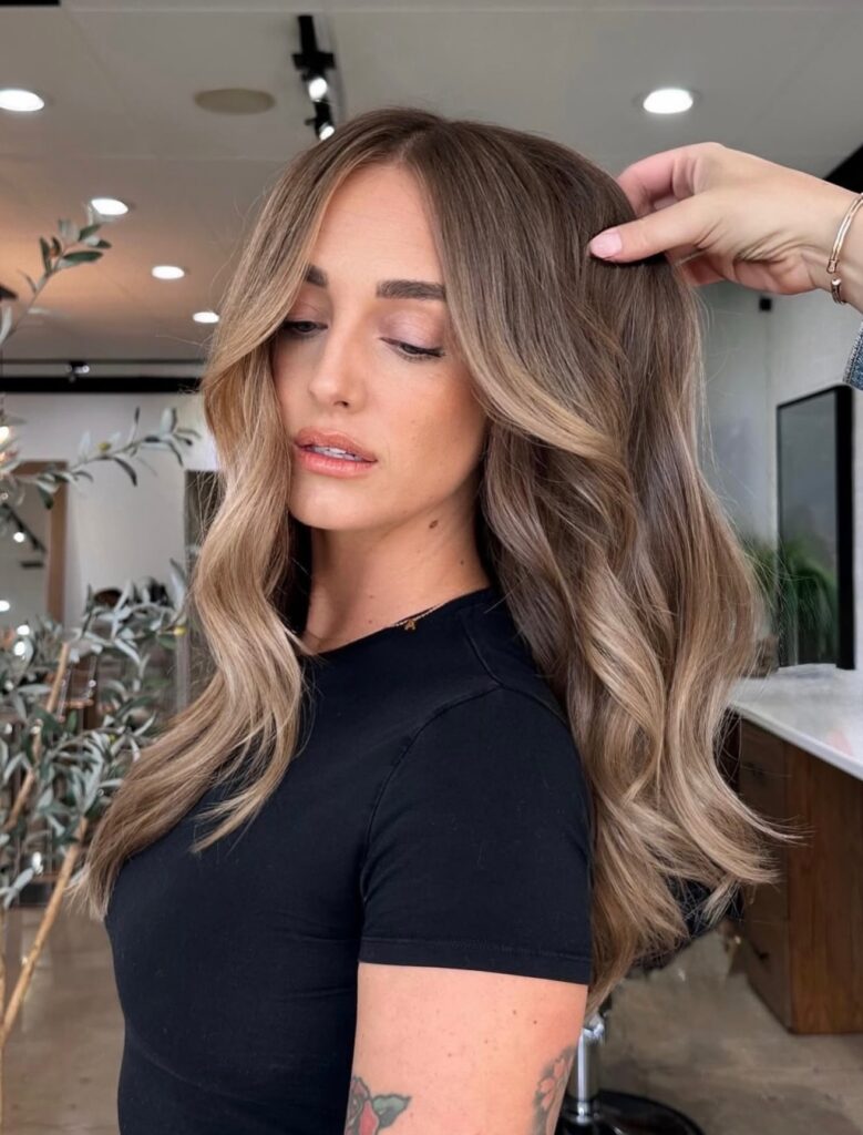 A woman with long mushroom brown hair and light sandy blonde highlights styled in voluminous face framing waves.