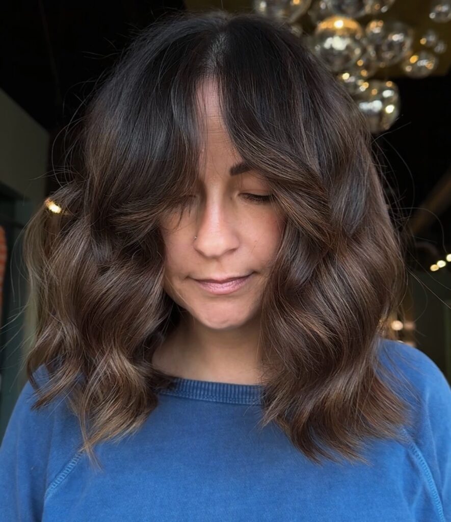 A woman showcasing a voluminous mocha brown lob with wavy layers and soft curtain bangs.