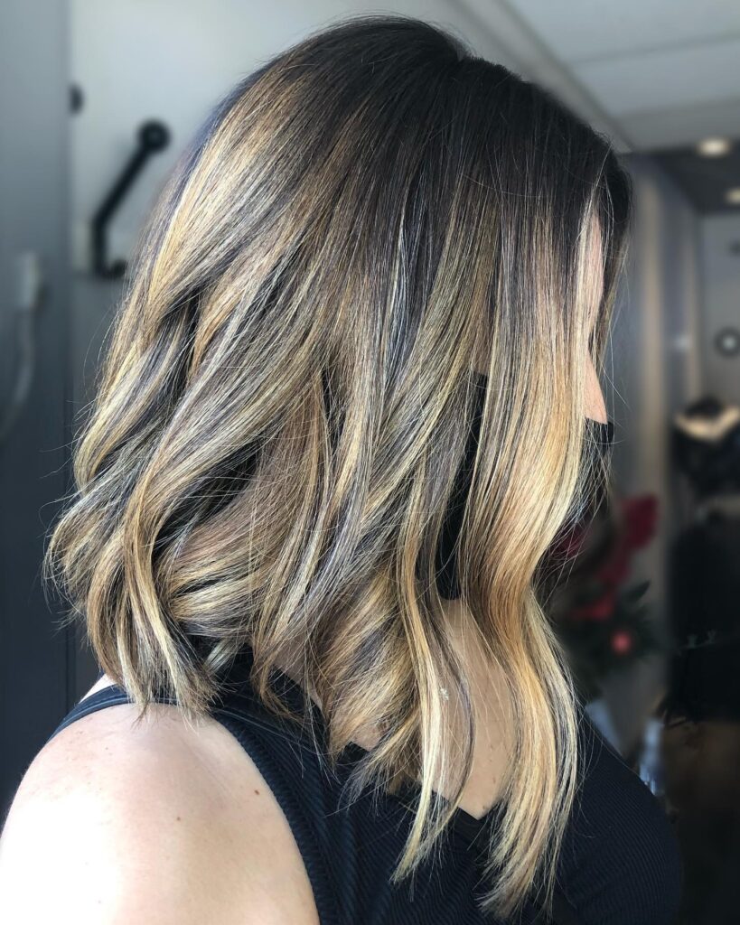 A side view of a woman with a dark brunette lob featuring chunky caramel balayage highlights and soft beachy waves.