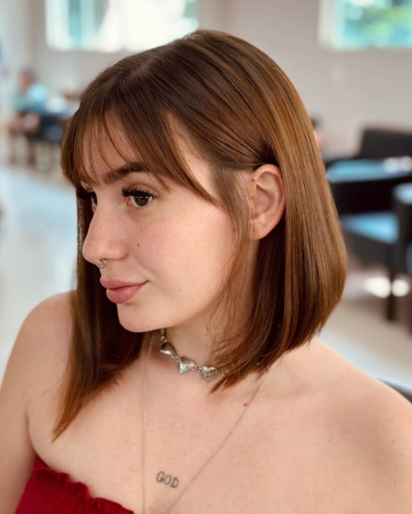 A young woman wearing a shoulder length auburn lob with soft wispy bangs and subtle curving ends.